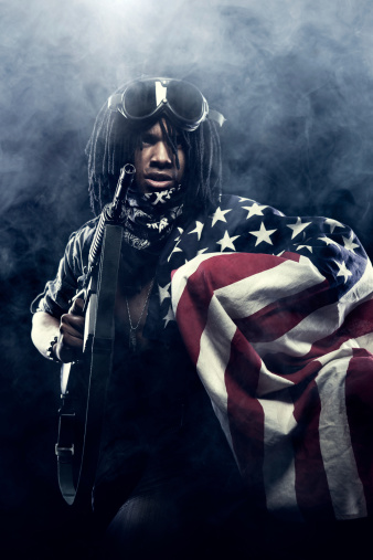 An African American man with dreadlocks and a bandanna mask holds an M16 machine gun and a worn American flag in a smoke filled battle.  Vertical with copy space.