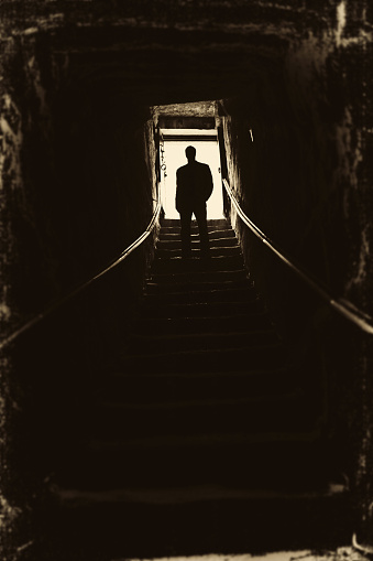 An elderly man in a hat and coat walks up the stairs in the fog under a black umbrella. Lonely sad man in scarf with umbrella walking under rain. Back view of an old man. Sadness, longing, loneliness.