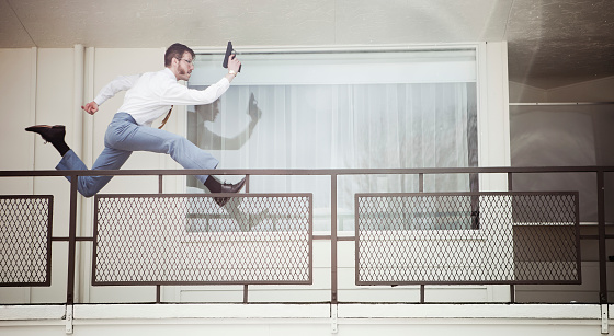 A two dimensional view of a spy or undercover agent running down a vintage California style hotel / motel balcony, gun in hand.  Big chops sideburns, classy mustache, and glasses. Horizontal with copy space; LOW CONTRAST intentional to emulate a 1970's style film look.   SLIGHT MOTION BLUR.