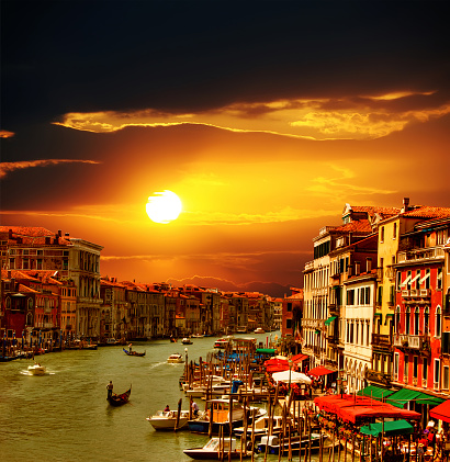 Beautiful view of Grand Canal at sunset in Venice, visible are boats, gondolas, restaurants, coffee shops and Venetian traditional architecture. Magnificent sun and clouds in the background. Venice, Italy.