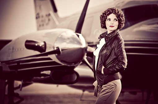 Horizontal portrait of a young woman dressed in 40's aviator attire, standing in front of her plane looking back with a slight smile on her face.
