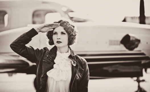 Horizontal portrait of a young woman dressed in 40's aviator attire, standing in front of her plane saluting. Image is processed to look vintage and has a slight grain effect.