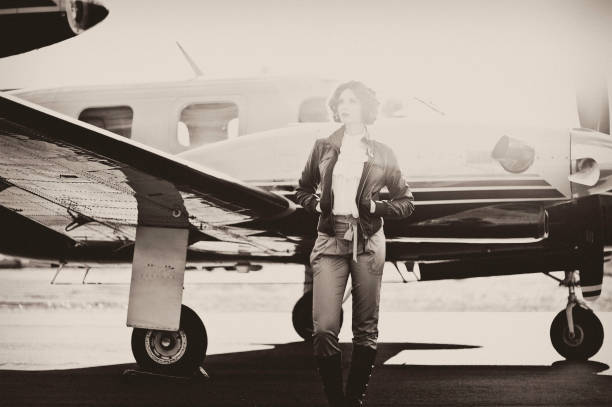 Horizontal portrait of a young woman dressed in 40's aviator attire, standing in front of her plane, looking out with a sun flare behind her. Image is processed to look vintage and has a slight grain effect.