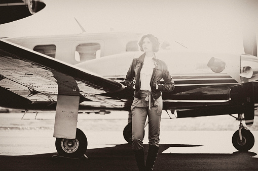 Horizontal portrait of a young woman dressed in 40's aviator attire, standing in front of her plane, looking out with a sun flare behind her. Image is processed to look vintage and has a slight grain effect.