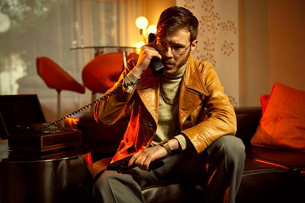 A serious 1970's style undercover spy or mobster sporting a leather jacket, turtleneck, gold chain and slacks, sits on a leather couch with a pistol in a vintage styled room, talking on a phone that closes into a wooden box.  Ransom call?  Heated Interrogation? Orange lighting.  Horizontal with copy space.