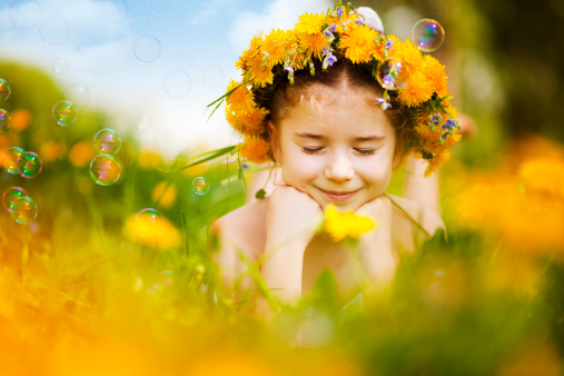 Cute little girl wearing dandelion wreath lying on flower meadow and smiling