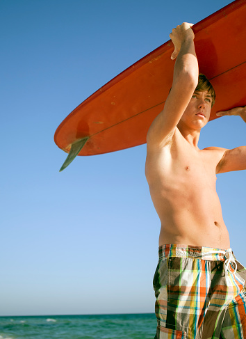 Teenage kid looking away while holding a red surf board above his head. Vertical shot.