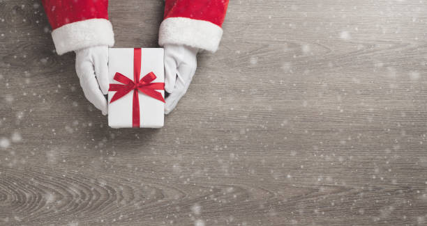 Santa claus hands is holding a white gift box with red ribbon Merry Christmas concept, Top view of Santa claus hands is holding a white gift box with red ribbon and snow on wooden background. Banner, Panoramic view for web. Copy space. holidays and seasonal stock pictures, royalty-free photos & images
