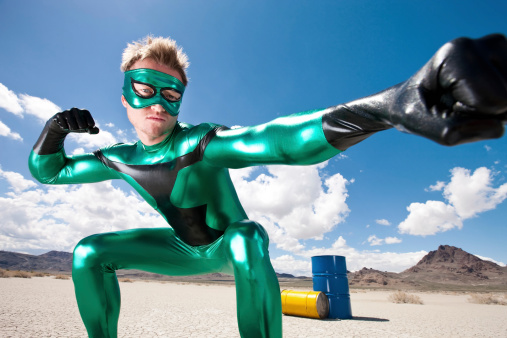 Low angle view of a masked hero in metallic-green costume ready to level kapow punch in defense of the environment against toxic waste disposal efforts in desert. Blue and yellow 50 gallon drums in background.