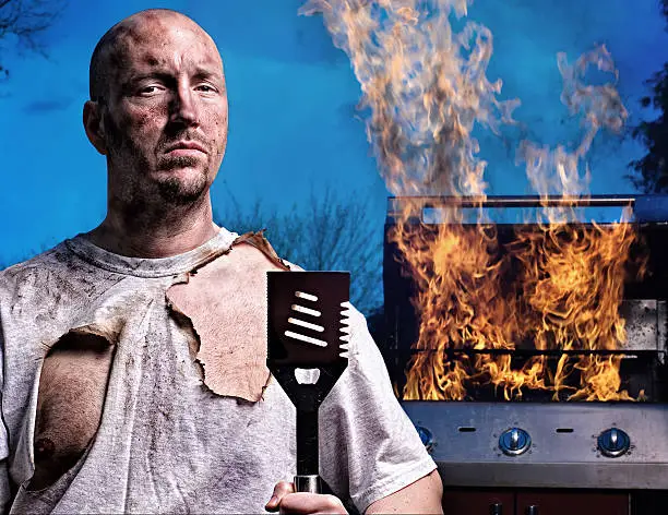 Man with burned face and shirt holding spatula in front of a flaming barbecue grill.