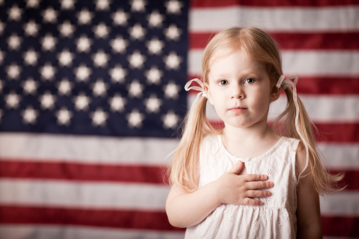 Color photo of a little blond-haired girl with her hand on her heart in front of an American flag.