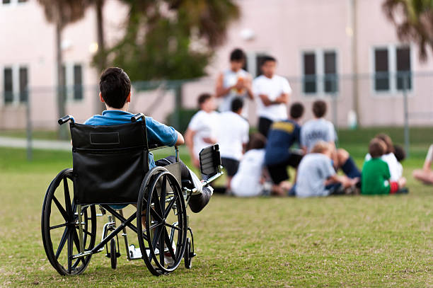 young disabled boy looking upon his peers leaving him out twelve years old child sitting in a wheel chair watching other kids getting together in the park while he is left behind prejudice stock pictures, royalty-free photos & images