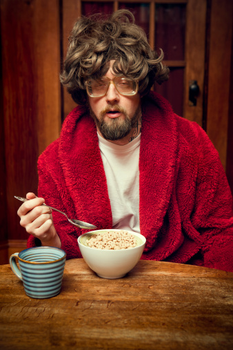 Not a morning person... A groggy, tired looking man with bad hair, nerdy glasses, and a bathrobe looks like he is about to fall asleep while eating his bowl of breakfast cereal.  Stained hardwood paneling and table. Vertical with copy space.