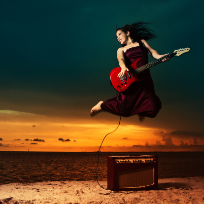 teenager jumping on the air and playing an electric guitar in the beach wearing a long red dress at sunset