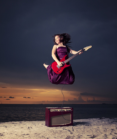 teenager jumping on the air and playing an electric guitar in the beach wearing a long red dress at sunset