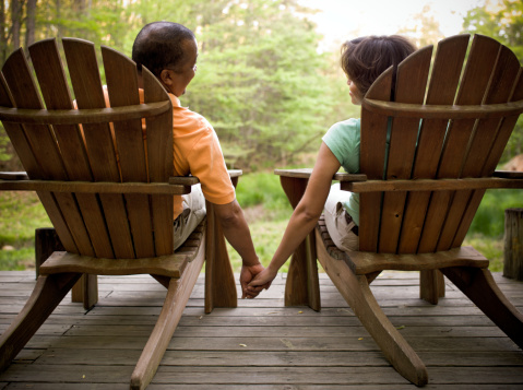 Attractive ethnic couple holding hands and sitting in Adirondack chairs looking over a meadow. Horizontal shot.