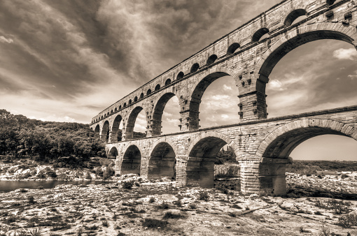 The photo is  HDR (high dynamic range) obtained by combining several images with different exposure time. The photo was then postprocessed by converting to B/W and toning. The Pont du Gard is a notable ancient Roman aqueduct bridge that crosses the Gard River in southern France. It is part of a 50 km (31 mi) long aqueduct that runs between Uzes and Nimes in the South of France.