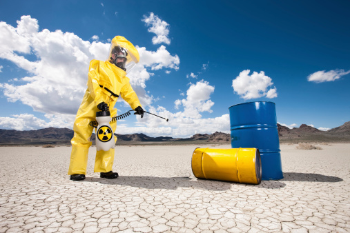 Man in hazardous materials protective gear using chemical neutralizing spray equipment in desert near 50-gal drums of chemical waste.