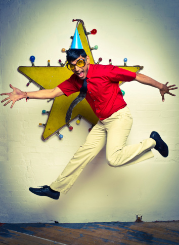 A young male adult wearing a party hat and novelty sunglasses jumps in front a wall decoration.