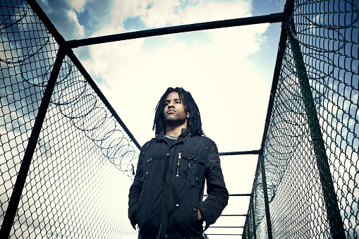A young African American man with dreadlocks stands with hands in pockets in between two barbed wire fence.  The sun rises in a vibrant blue sky behind him with small cloudscapes.  Horizontal with copy space.