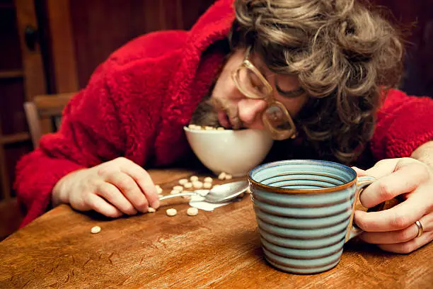 The coffee's not working... A groggy, tired looking man with bad hair, nerdy glasses, and a bathrobe has fallen asleep while eating his bowl of breakfast cereal, spilling milk and food on the table.  Stained hardwood paneling and table. Horizontal.