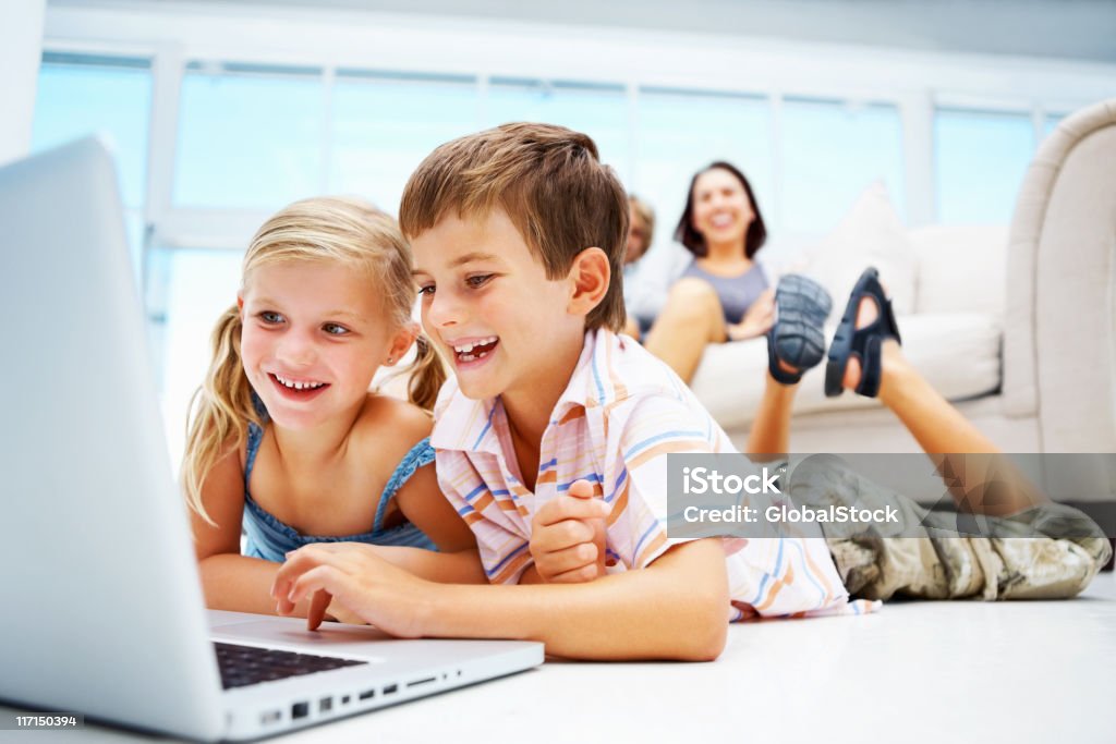 Kids using laptop on floor with parents in the background Portrait of young kids using laptop on floor while parents sitting on sofa in the background Adult Stock Photo