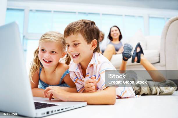 Kids Using Laptop On Floor With Parents In The Background Stock Photo - Download Image Now