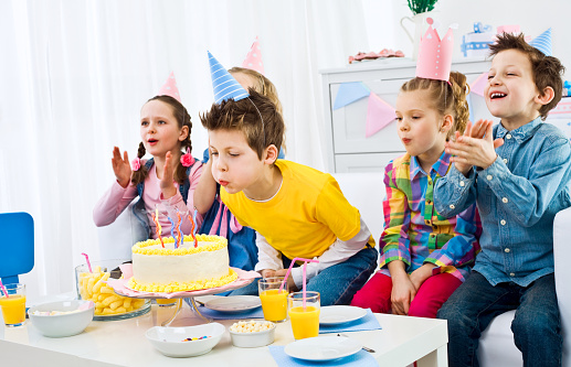 Five cute, smiling kids wearing party hats having fun at birthday party. The boy in yellow t-shirt, sitting in the middle blowing out birthday candles.