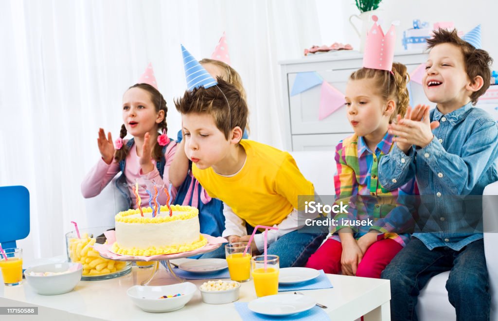 Kids birthday party Five cute, smiling kids wearing party hats having fun at birthday party. The boy in yellow t-shirt, sitting in the middle blowing out birthday candles. Birthday Candle Stock Photo