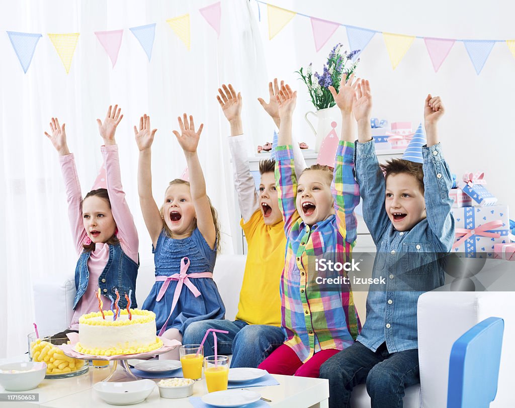 Happy birthday! Birthday Party. Five happy kids sitting in a row on sofa at home and raising arms in celebration, looking away and yelling. Birthday cake in front of them on the table. 6-7 Years Stock Photo