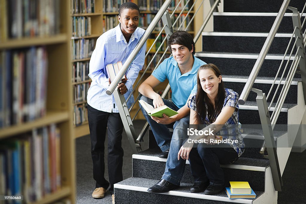 Tre sorridenti studenti raccogliere all'interno di una libreria - Foto stock royalty-free di Abbigliamento casual