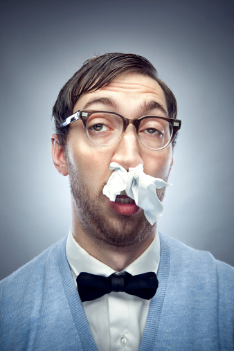 Nerdy IT young man wearing a bow tie, blue sweater vest, and horn rimmed glasses makes a funny sad face, a kleenex tissue stuffed in his nostrils to stop his cold /allergies.   Vertical on gray / blue background.