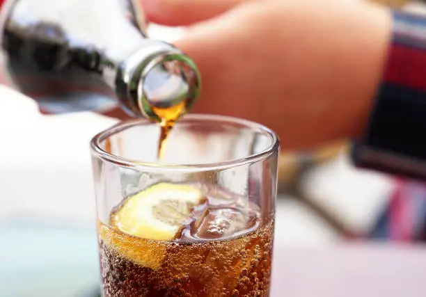 Photo of Hand pouring cola soda drink from bottle to glass with ice cubes and lemon slices