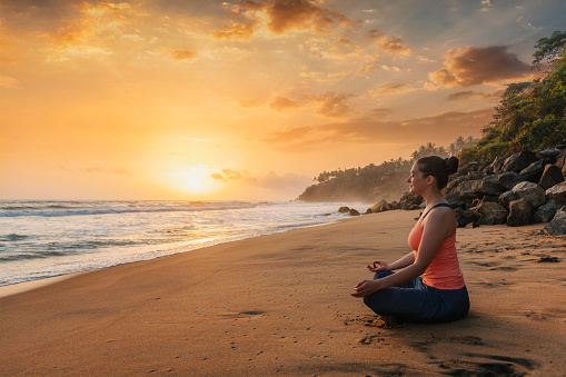 Woman doing yoga - meditate and relax in Padmasana Lotus asana pose with chin mudra outdoors at tropical beach on sunset with dramatic sun