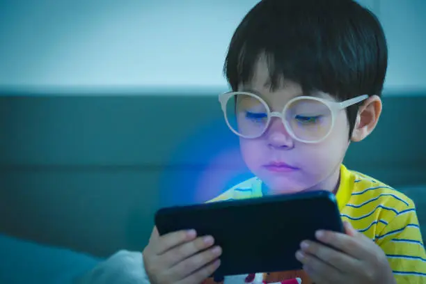 Cute little child watching cartoon on smartphone in the dark room. Dangers of blue light damage his eyes. Handsome little boy can get age related macular degeneration of blue light, he wear eyeglasses