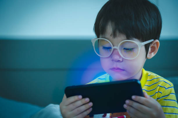 симпатичный маленький ребенок смотрит мультфильм на смартфоне в темной комнате. опасности синего света повреждают глаза. красивый маленьк - glasses child little boys happiness стоковые фото и изображения