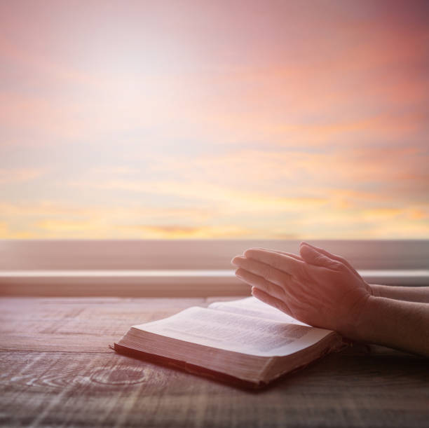 istock Close up of woman's praying hands reading Bible, with dramatic light. Wood table with sun rays coming through window. Christian image 1171533449