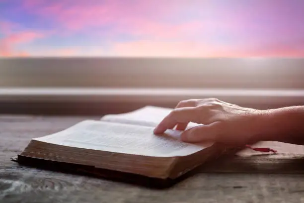 Close up of woman's hand reading Bible, with dramatic light. Wood table with sun rays coming through window. Christian image