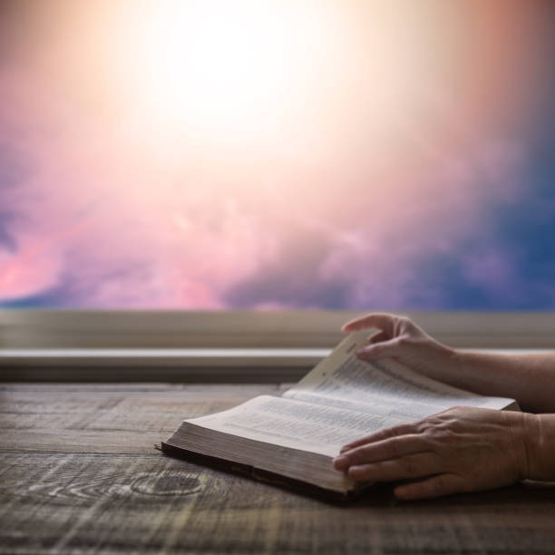 close up of woman's hand reading bible, turning page, with dramatic light. wood table with sun rays coming through window. christian image - praying human hand worshipper wood imagens e fotografias de stock