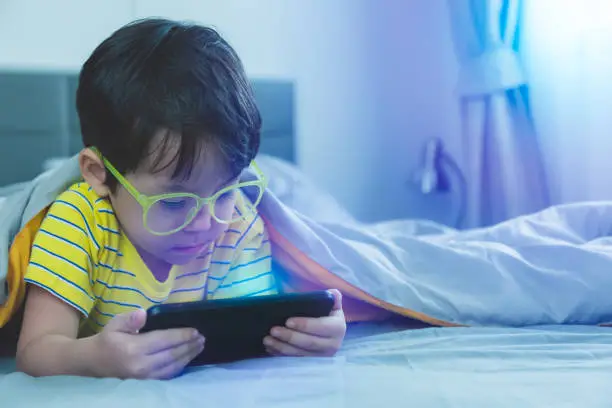 Photo of Cute little child watch movie on smartphone at bed. Dangers of blue light can damage eyes. Handsome little boy can be age related macular degeneration from blue light, wear eyeglasses since childhood