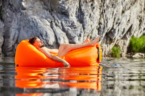Man sleeping in a raft on the river. The tourist fell asleep in an orange air sofa or an inflatable lounge floating on the river. Rafting on the wild river on an inflatable lounger, while on vacation, ecotourism. chaise longue stock pictures, royalty-free photos & images