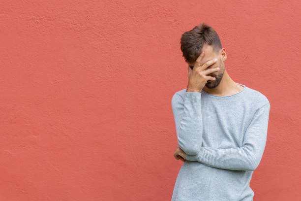 Facepalm. Ashamed embarrassed man covering his face. Young guy on red wall background with copy space. Emotion facial expression and feelings concept