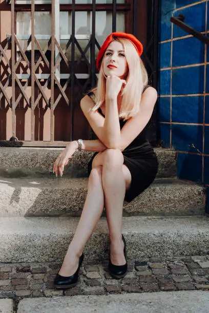 Stylish young woman in a red beret, black dress and high heels posing seated on exterior steps in town with her hand to her chin and a thoughtful expression