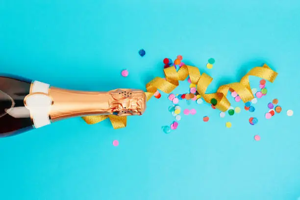 Bottle of champagne, golden ribbon and colored confetti over blue background. Flat lay, top view