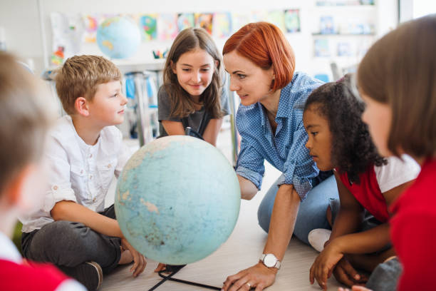 группа маленьких школьников с учителем, сидящим на полу в классе, учится. - learning child education globe стоковые фото и изображения
