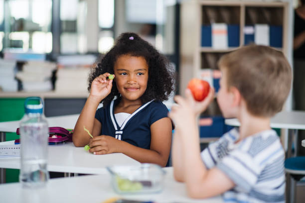 small school children sitting at the desk in classroom, eating fruit. - child food school children eating imagens e fotografias de stock