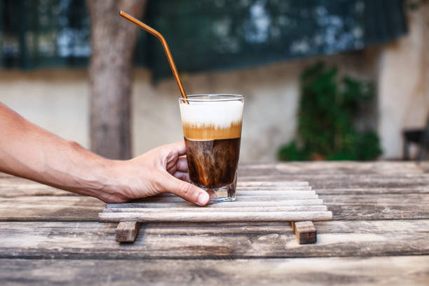 A hand holding an ice coffee with milk and a stainless steel straw on a wooden table A hand holding an ice coffee with milk and a stainless steel straw on a wooden table freddo cappuccino stock pictures, royalty-free photos & images