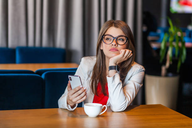 portrait of serious thoughtful beautiful stylish brunette young woman in glasses sitting, holding her mobile smart phone and thinking about something. - pensive question mark teenager adversity imagens e fotografias de stock