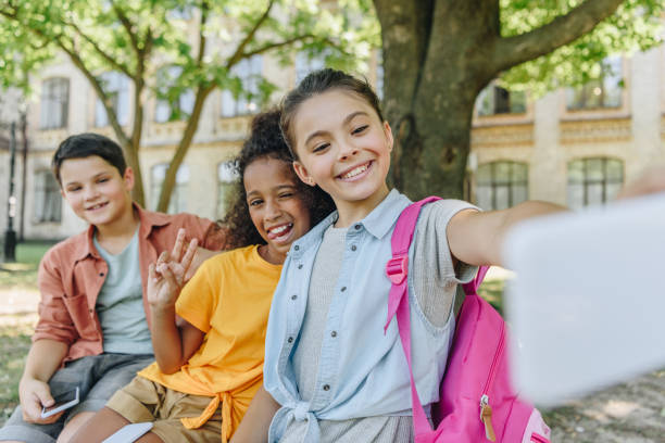 istock cheerful schoolgirl taking selfie with multicultural friends while sitting on bench in schoolyard 1171585945