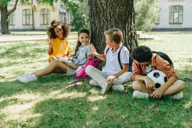 four cute multicultural schoolkids talking while sitting on lawn under tree